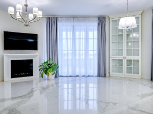 Bright living room with marble floor, white fireplace and wall-mounted TV, a potted plant, and a glass-front cabinet by sheer-curtained windowsill light.